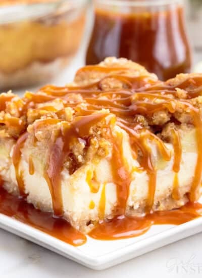 A square of apple cheesecake bar served with caramel sauce topping on a small plate, jar of caramel sauce.