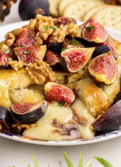 Close up of baked brie with fig jam, topped with quartered figs, chopped candied walnuts, and fresh herbs. Crackers and other fresh ingredients scattered around in the background.