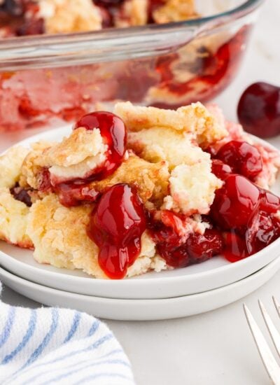 A serving of Cherry Cheesecake Dump Cake on small white plate, with linen, forks, and baking dish nearby..