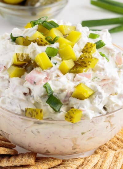 Bowl of dill pickle dip with ham and chopped pickles on top, on a tray with crackers.