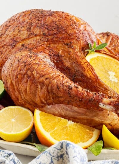 Fully cooked turkey from this easy smoked turkey recipe on a platter garnished with fresh orange and lemon slices and fresh herbs.