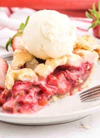 Slice of strawberry rhubarb pie on a dessert plate, topped with scoop of vanilla ice cream and fresh strawberry halves.