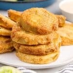 Stack of fried green tomatoes on a platter, being served with a fork, and a bowl of green tomatoes in the background.