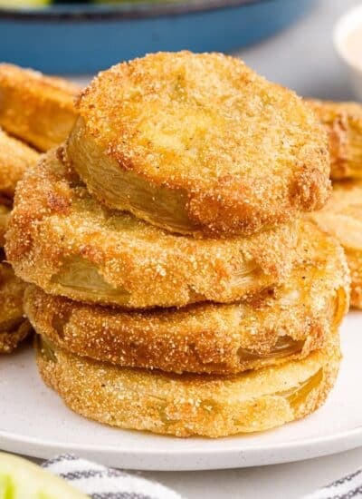 Stack of fried green tomatoes on a platter, being served with a fork, and a bowl of green tomatoes in the background.