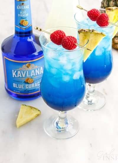 Close up of two tall glasses with fruit tingle cocktail and garnished with pineapple wedges and skewered raspberries, bottle of Blue Curacao, pineapple wedge garnish on a marble countertop.