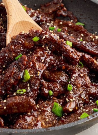 Skillet full of ginger beef garnished with green onions and sesame seeds, with wooden spoon scooping.