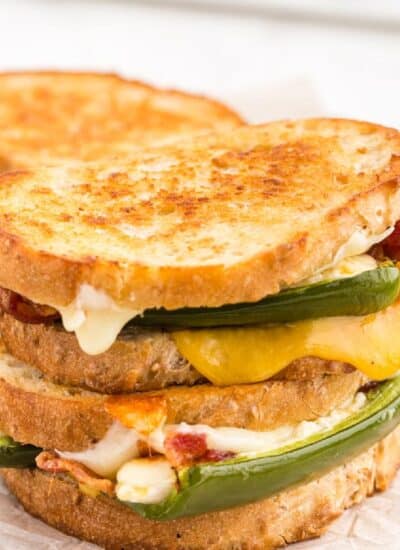 Two Jalape&ntilde;o popper grilled cheese sandwiches stacked on parchment paper-lined wooden kitchen board, stuffed jalapeno poppers, crispy bacon strips, on a white marble countertop.