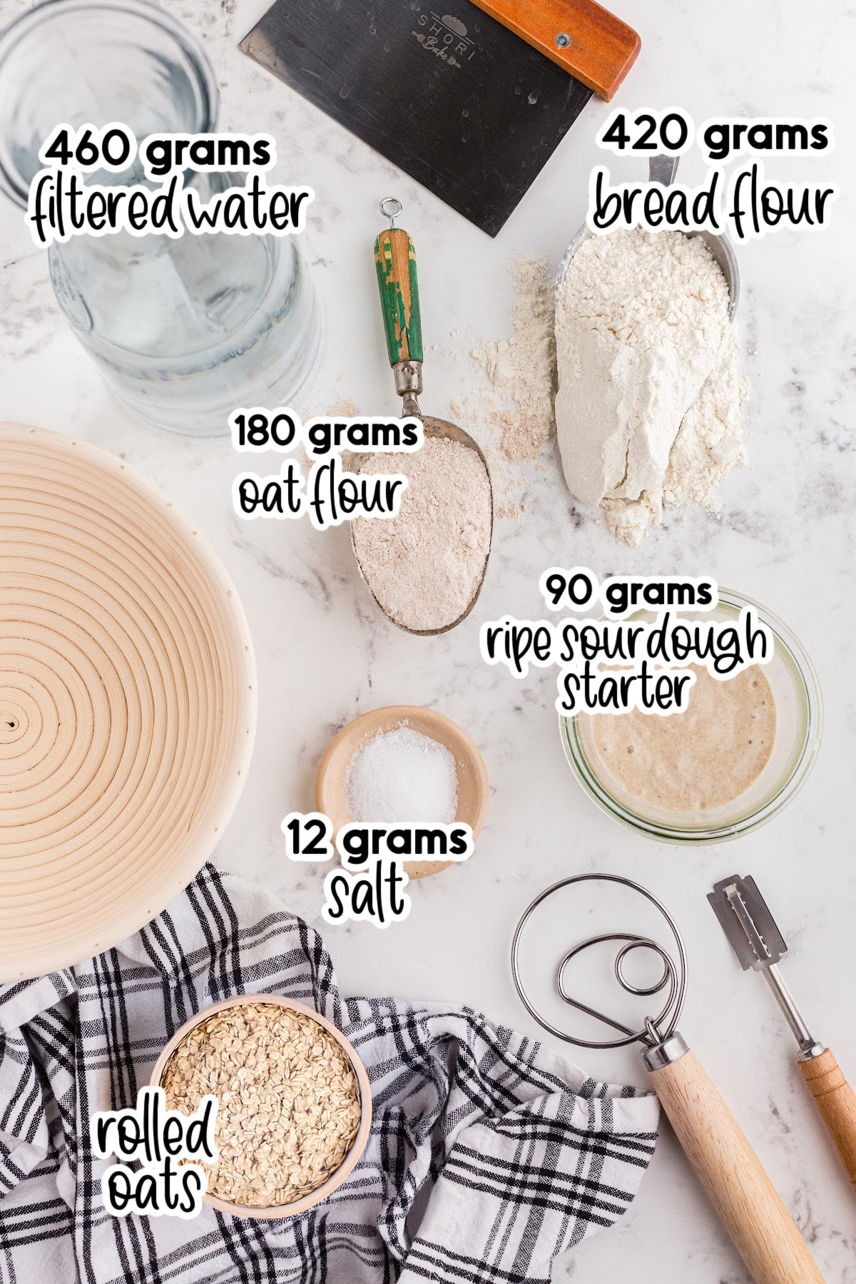 Ingredients and tools to make oat sourdough bread on a marble countertop with a navy and white checkered kitchen cloth.