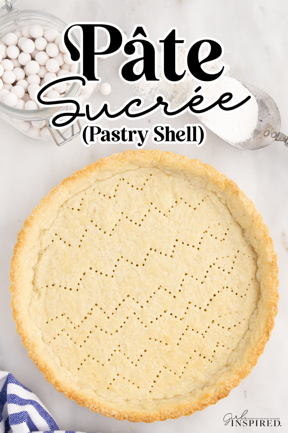 Overhead view of baked pate sucree shell on a white countertop, white and blue striped kitchen cloth, sugar scoop, and pie beads.