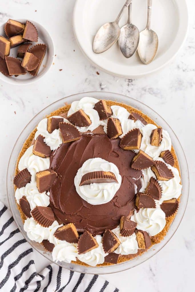 Glass pie dish with graham cracker crust filled with peanut butter chocolate ice cream decorated with whipped cream and chocolate ganache and cut peanut butter cups, grey and white striped linen, stack of white cake plates and teaspoons, bowl with cut peanut butter cups on marble counter top
