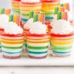 Rainbow Jello Shots garnished with whipped cream and rainbow candies on a white serving board.