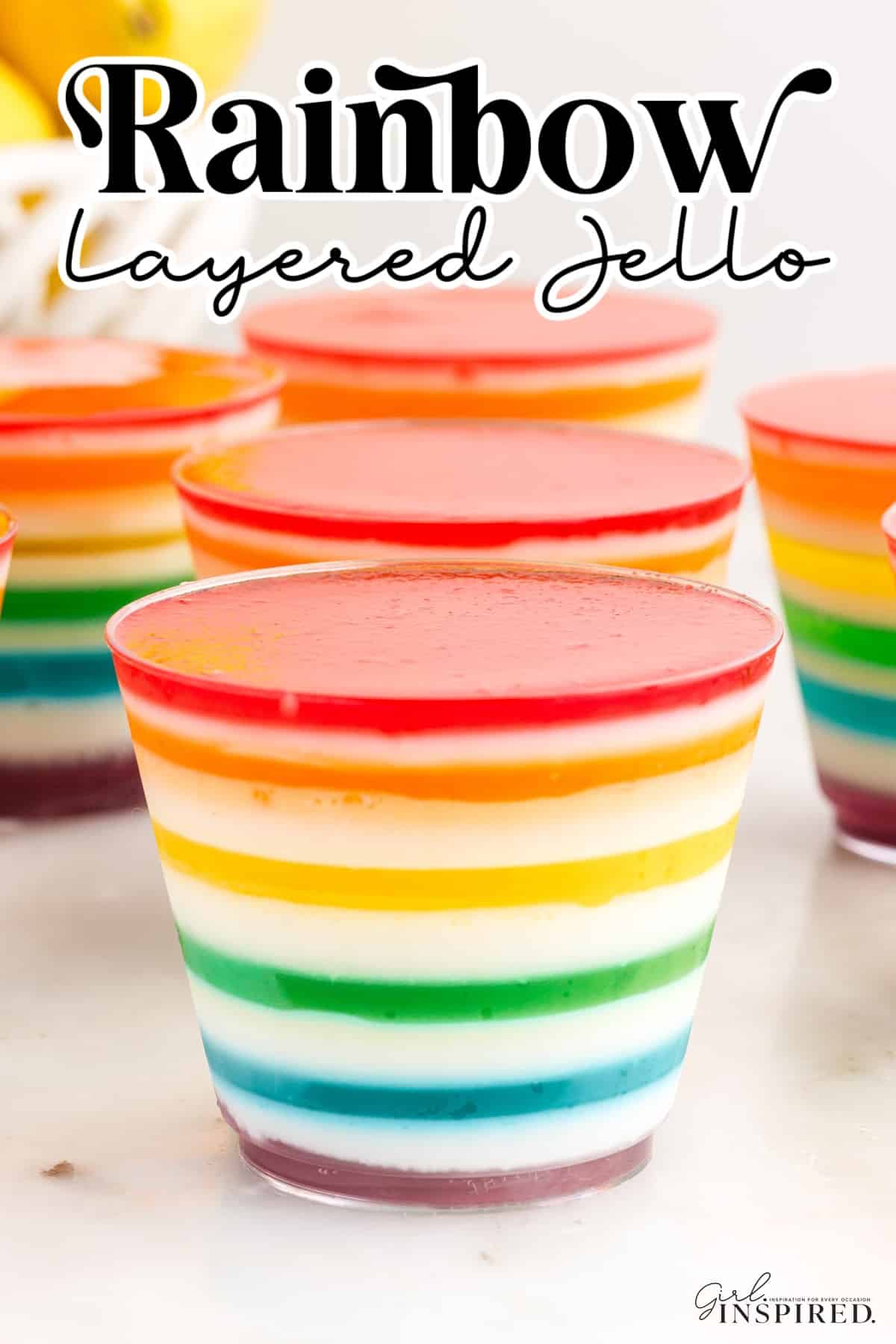 Rainbow layered jello cups on a marble countertop with a bowl of fresh lemons in the background.