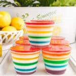 Rainbow layered jello cups on a white rimmed serving tray, a bowl of fresh lemons and bow of yellow jello in the background.