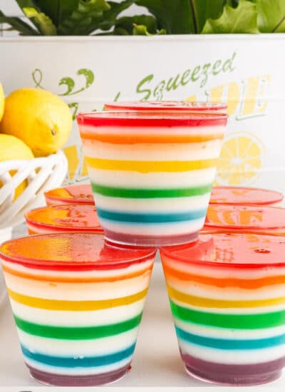 Rainbow layered jello cups on a white rimmed serving tray, a bowl of fresh lemons and bow of yellow jello in the background.