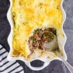 Overhead view of shepherd's pie with leftover roast beef, a single serving removed from the pie dish with a silver serving spoon, striped kitchen cloth alongside the pie dish on a dark marble countertop.