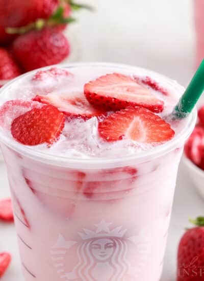 Pink drink recipe filling a plastic cup with fresh strawberries floating on top, bowl of strawberries, green straw.