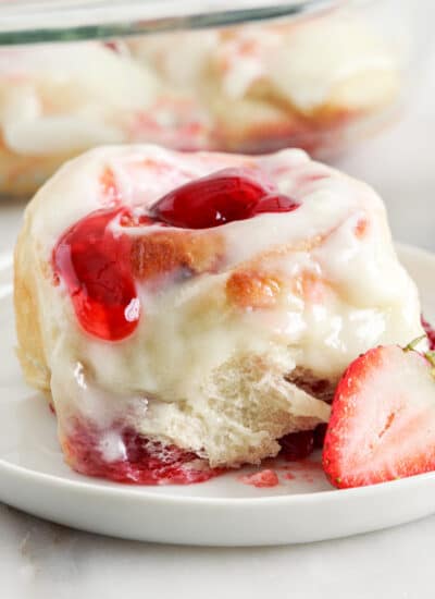 close up of a strawberry cinnamon roll, showing the frosting and sliced berries, with other cinnamon rolls in the background.