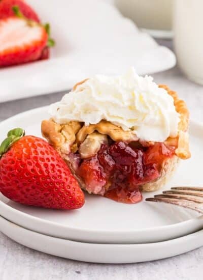 Strawberry Rhubarb mini pie on a plate, cut open to show the strawberry inside, topped with whipped cream and a whole strawberry on the plate, with other mini pies in the background., with text overlay.